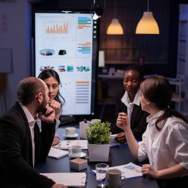 Workaholics businesspeople brainstorming financial company ideas analyzing strategy paperwork late at night in business office meeting room. Multi-ethnic coworkers checking solution